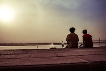 a couple sitting on a concrete wall by a shore at sunset 