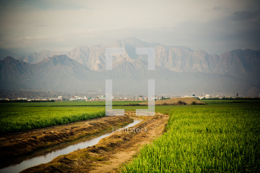 Stream running through a grass field with a city and mountains in the background