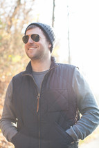 smiling man standing outdoors in sunglasses and a wool cap 
