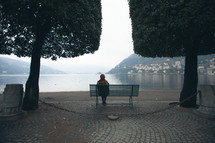 a woman sitting on a park bench looking out at a bay under fog 