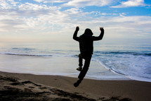Silhouette of a person in a hoodie jumping for joy on the beach near the ocean.