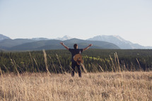 a man with a guitar standing in a field with open arms and a mountain view 