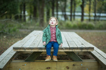 a toddler boy outdoors in a jacket sitting on a picnic table 