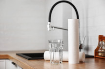 A glass of clean water and foamed polypropylene filter cartridges on wooden table in a kitchen interior. Installation of reverse osmosis water purification system. Concept Household filtration system