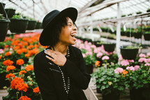An African American woman in a black hat standing in a green house full of flowers 