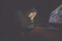 A tired man in a hooded sweatshirt in front of a laptop computer.