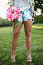 the legs of a young woman in shorts holding a flower and sunglasses 