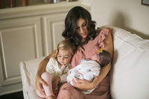 mother holding a swaddled newborn baby and toddler daughter on a chair 