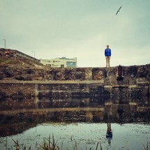 Man standing on rocks at edge of water with hill and building and flying bird in the background.