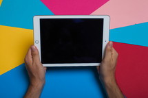 Hands holding an electronic tablet on a multi-colored background.