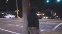 a young man leaning against a pole on a city sidewalk at night 