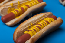 Hot dogs and buns drizzled with mustard on a blue background.