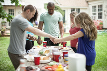 friends holding hands in prayer over an outdoor table before eating 