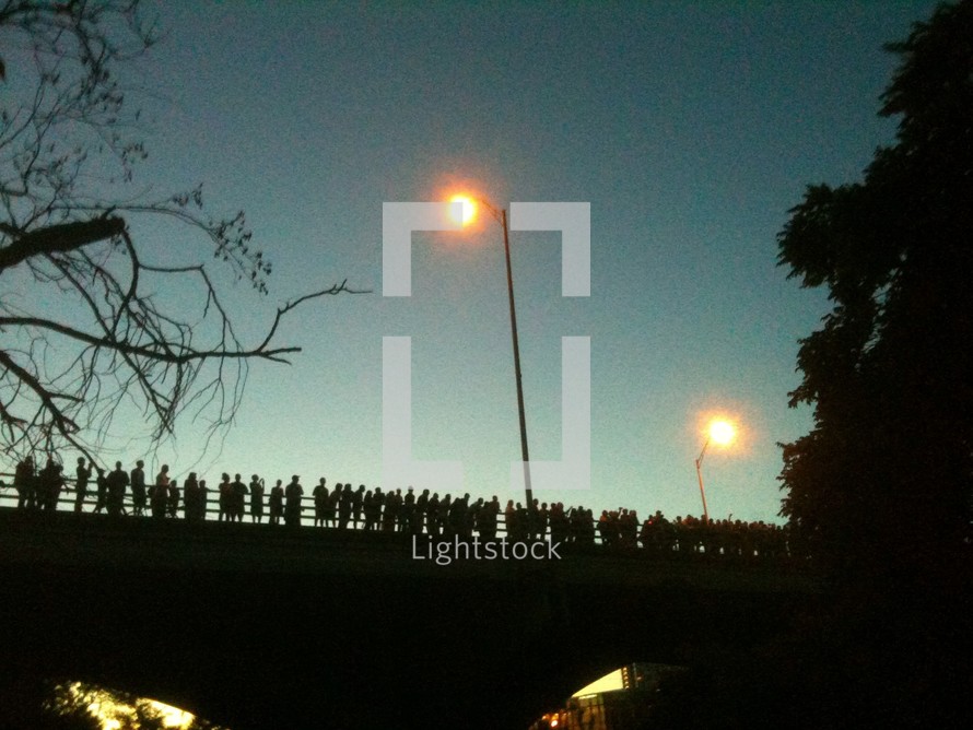 Silhouette of crowd of people standing on bridge with streetlights at dusk.