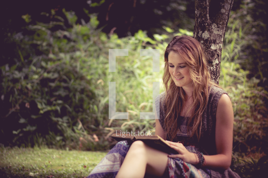 A young woman sitting against a tree and reading a book.