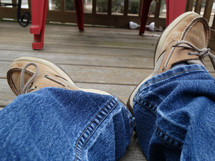 a person's legs laying on porch deck wearing jeans and brown boat shoes with red decor in background