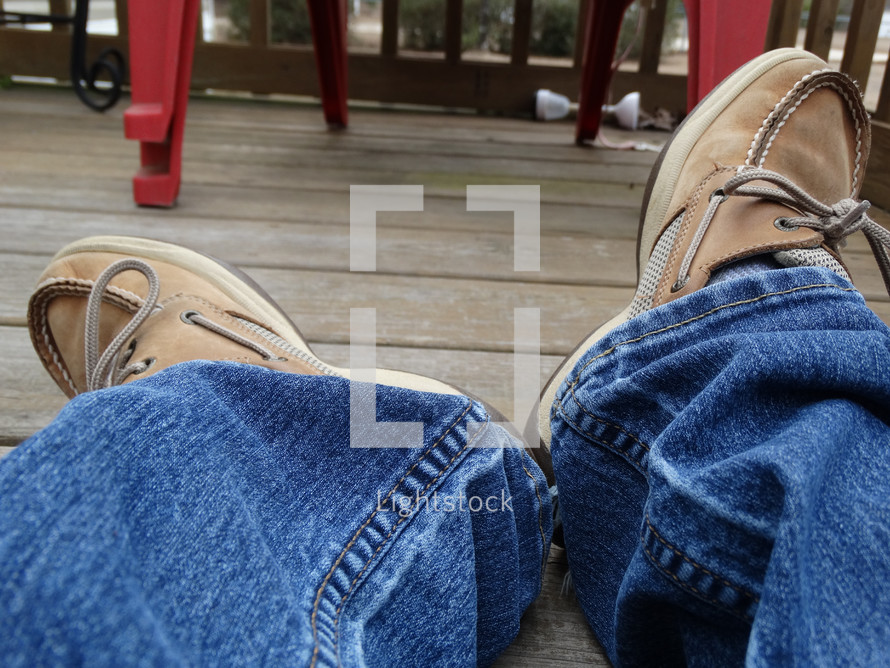 a person's legs laying on porch deck wearing jeans and brown boat shoes with red decor in background