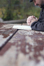 man praying near an open Bible on a picnic table 