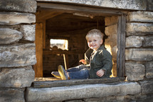 A toddler boy sitting in a window frame. 