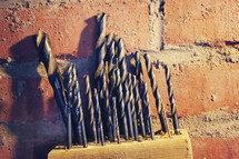 Drill bits in a block of wood against a brick wall.