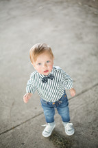 Toddler boy in a bow tie standing on the sidewalk outside.
