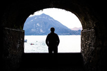 silhouette of a man standing in a tunnel and looking out at a lake 