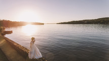 a bride standing by a lake shore at sunset 