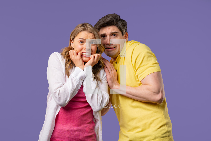 Frightened white american couple afraid of something and looks into camera with big eyes full of horror on violet wall background. Blonde woman and man. High quality 