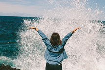 Woman with her arms raised as she faces the splash of the ocean water.