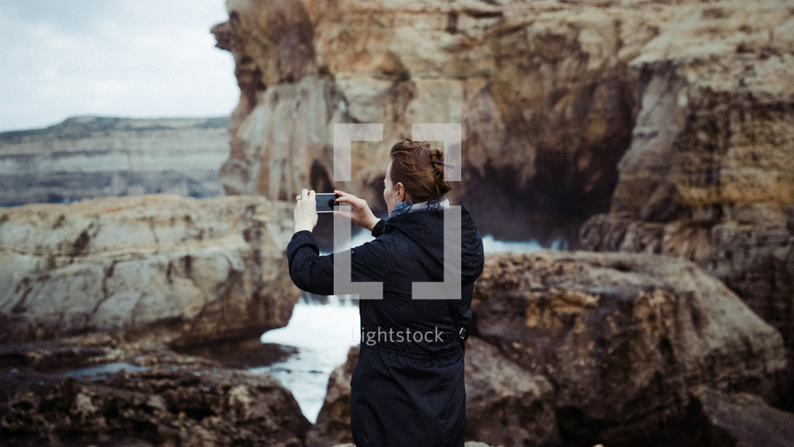 a woman taking a picture of waves crashing into rocks with her phone 