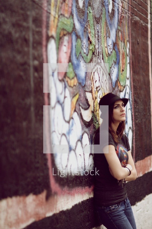 A teenage girl leans up against a brick graffiti wall with arms crossed