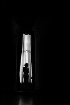 Silhouette of a woman in front of a tall curtained window.