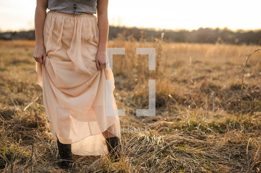 a woman in a long skirt and cowboy boots standing in dry grasses 