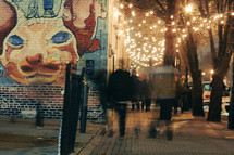 pedestrians walking down a city sidewalk at night and cat mural 