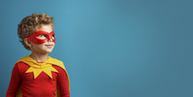 Cheerful young boy in red superhero costume with mask, looking up with inspiration against a teal background.