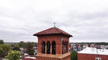 Circling a church tower with a cross on top on a cloudy overcast day with a neighborhood in the background.