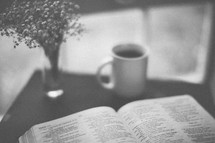 An open Bible on a table and a coffee cup and flower vase 