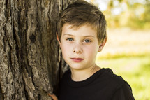 a boy child leaning against a tree trunk 