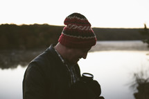 man in a wool cap standing by a lake holding a camera 