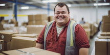 Happy male worker with Down syndrome in a red shirt and reflective vest, packaging boxes in a warehouse, with a bright smile and joyful expression.