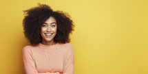 Joyful young woman with curly hair in a peach sweater, radiating happiness against a bright yellow background.