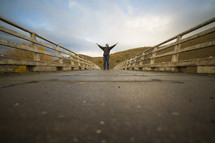 man standing on a bridge with hands raised to God 
