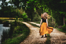 Girl with long hair in flowing skirt joyously twirling around with freedom in a country landscape with dirt road and pond.