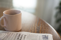 Morning light shining on a cup of coffee next to a newspaper on a table.