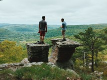 Two men stand on rock pinnacles overlooking a valley and forested hills.