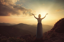 Woman on a hillside with arms raised in praise at daybreak.