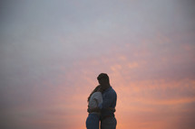 A man and woman embrace in front of a sunset.