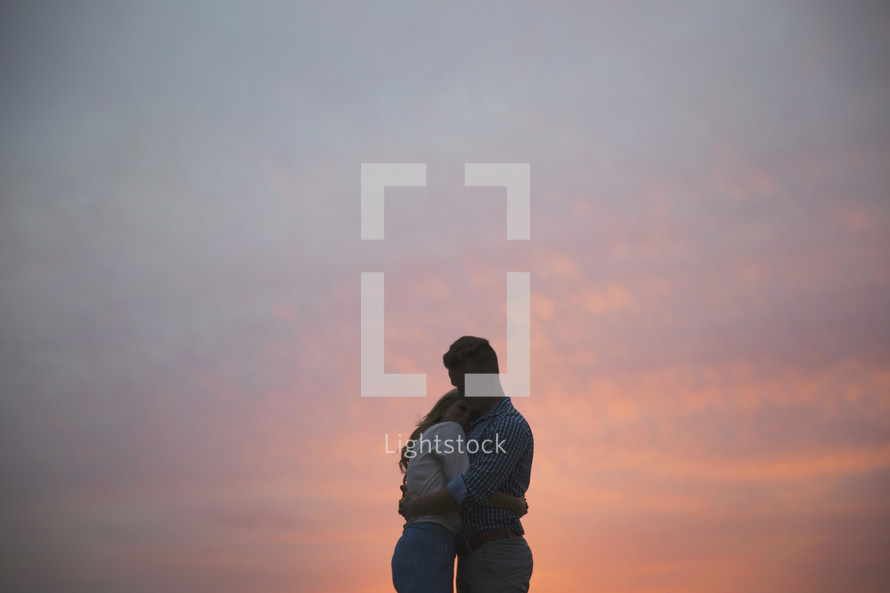 A man and woman embrace in front of a sunset.