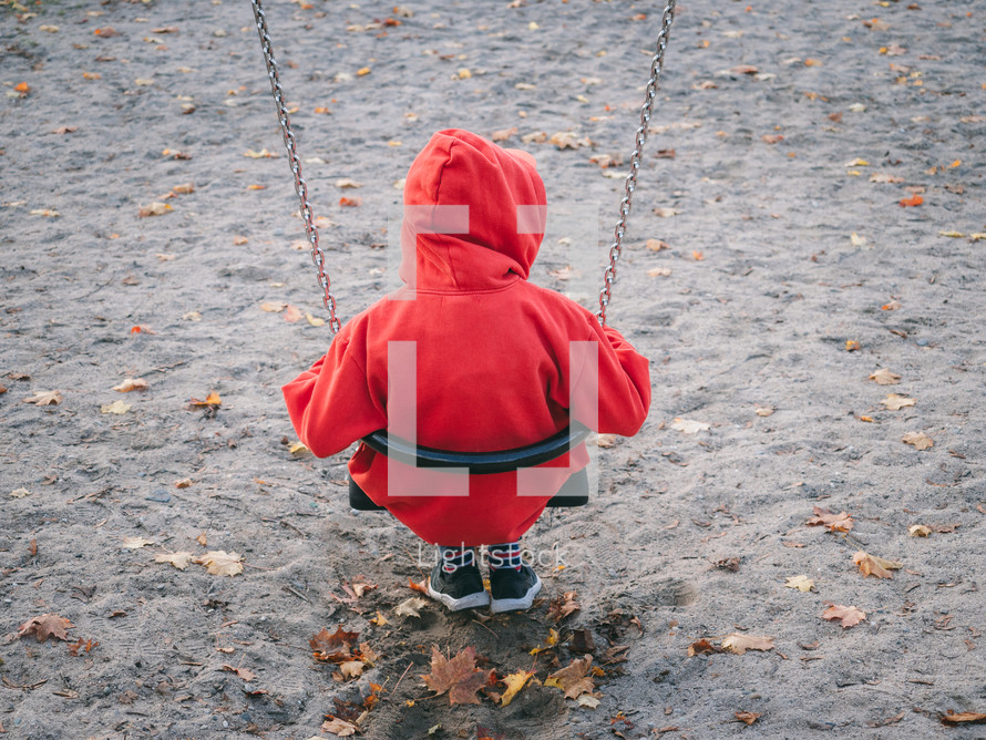 A child in a red hoodie on a sand-covered playground swing looking lonely into the distance