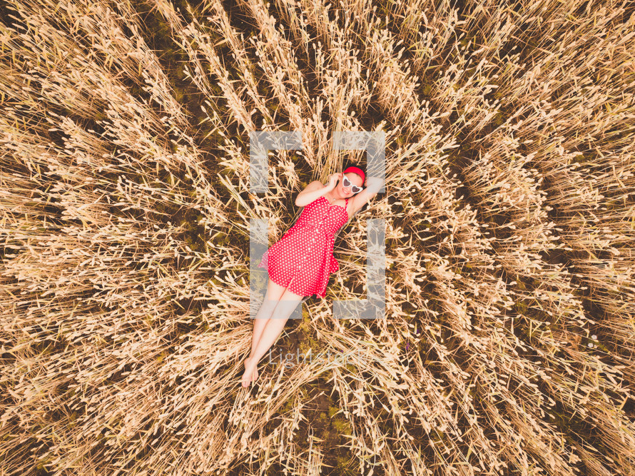  Young beautiful woman in red retro dress and sunglasses lying in wheat yellow field. Flying close above cornfield. AERIAL Drone view. Harvest, agriculture concept.
